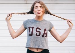 A woman playfully holds her braided hair out to the sides while making a kissy face, rocking a gray crop top with "USA" printed on it—perfect for anyone looking to shop for trendy women's T-shirts. She poses against a light-colored, horizontal-striped background.