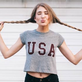 A woman playfully holds her braided hair out to the sides while making a kissy face, rocking a gray crop top with "USA" printed on it—perfect for anyone looking to shop for trendy women's T-shirts. She poses against a light-colored, horizontal-striped background.