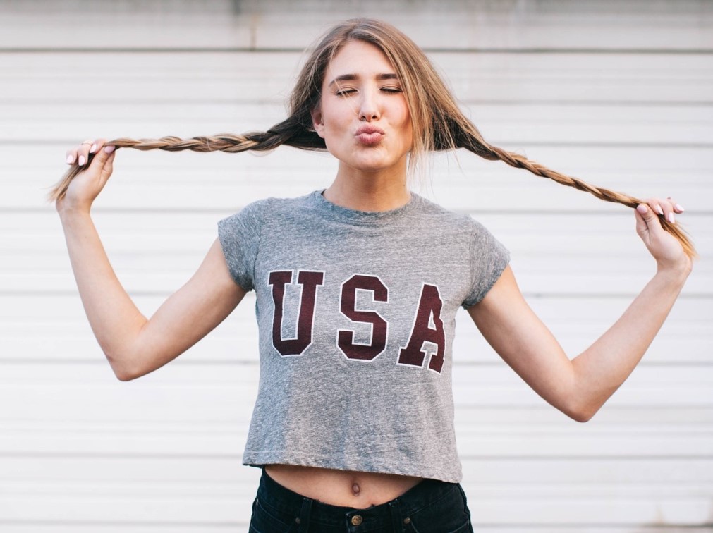 A woman playfully holds her braided hair out to the sides while making a kissy face, rocking a gray crop top with "USA" printed on it—perfect for anyone looking to shop for trendy women's T-shirts. She poses against a light-colored, horizontal-striped background.