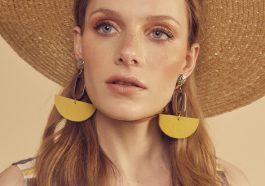 A woman with long, wavy hair, showcasing How To Look More Stylish, wears a wide-brimmed straw hat and large yellow half-circle earrings. Dressed in a sleeveless checkered top, she gazes slightly upward against a soft beige background.