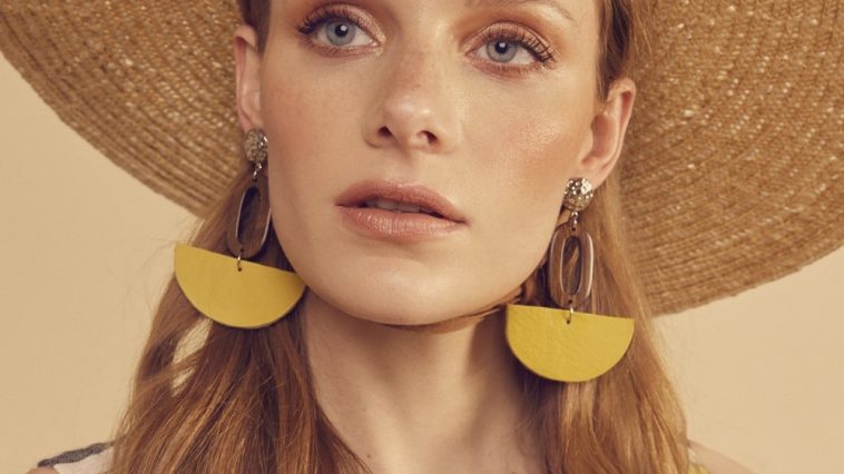 A woman with long, wavy hair, showcasing How To Look More Stylish, wears a wide-brimmed straw hat and large yellow half-circle earrings. Dressed in a sleeveless checkered top, she gazes slightly upward against a soft beige background.
