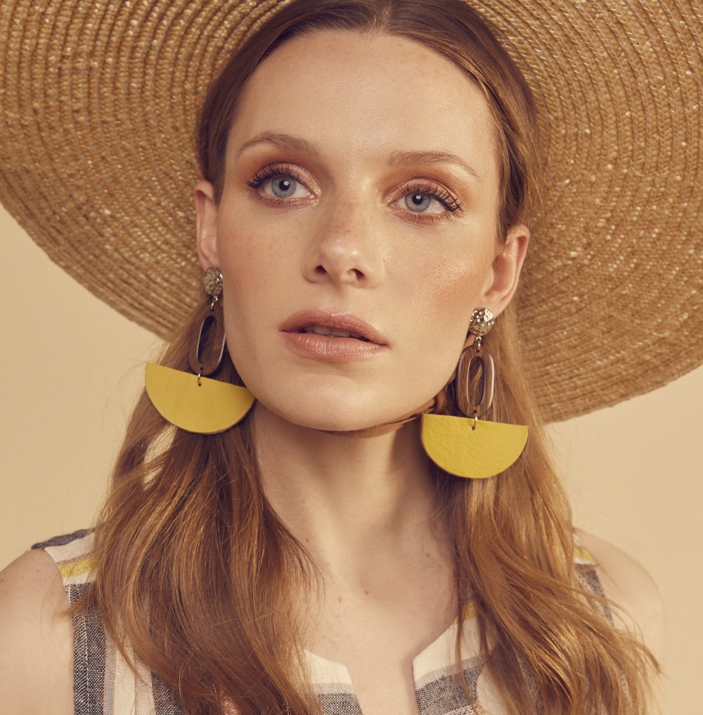 A woman with long, wavy hair, showcasing How To Look More Stylish, wears a wide-brimmed straw hat and large yellow half-circle earrings. Dressed in a sleeveless checkered top, she gazes slightly upward against a soft beige background.