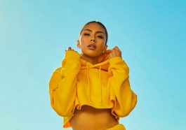 A person wearing a bright yellow cropped hoodie, their power color, stands against a clear blue sky. They have their hands raised towards the hood, looking directly at the camera. The outfit and backdrop create a vibrant and sunny atmosphere.