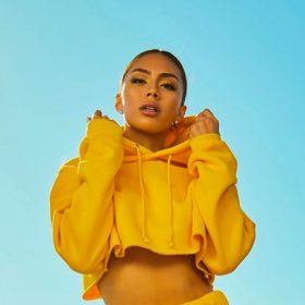 A person wearing a bright yellow cropped hoodie, their power color, stands against a clear blue sky. They have their hands raised towards the hood, looking directly at the camera. The outfit and backdrop create a vibrant and sunny atmosphere.