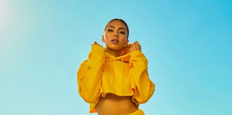 A person wearing a bright yellow cropped hoodie, their power color, stands against a clear blue sky. They have their hands raised towards the hood, looking directly at the camera. The outfit and backdrop create a vibrant and sunny atmosphere.